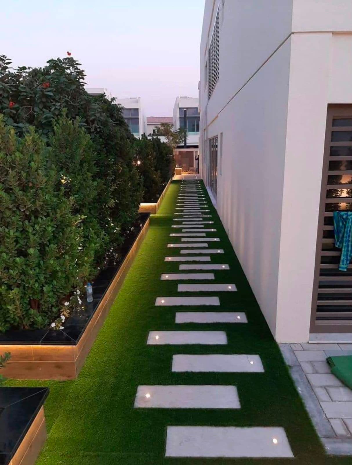 Side garden at dusk with stepping stones set into artificial grass, uplit planters and recessed ground lighting